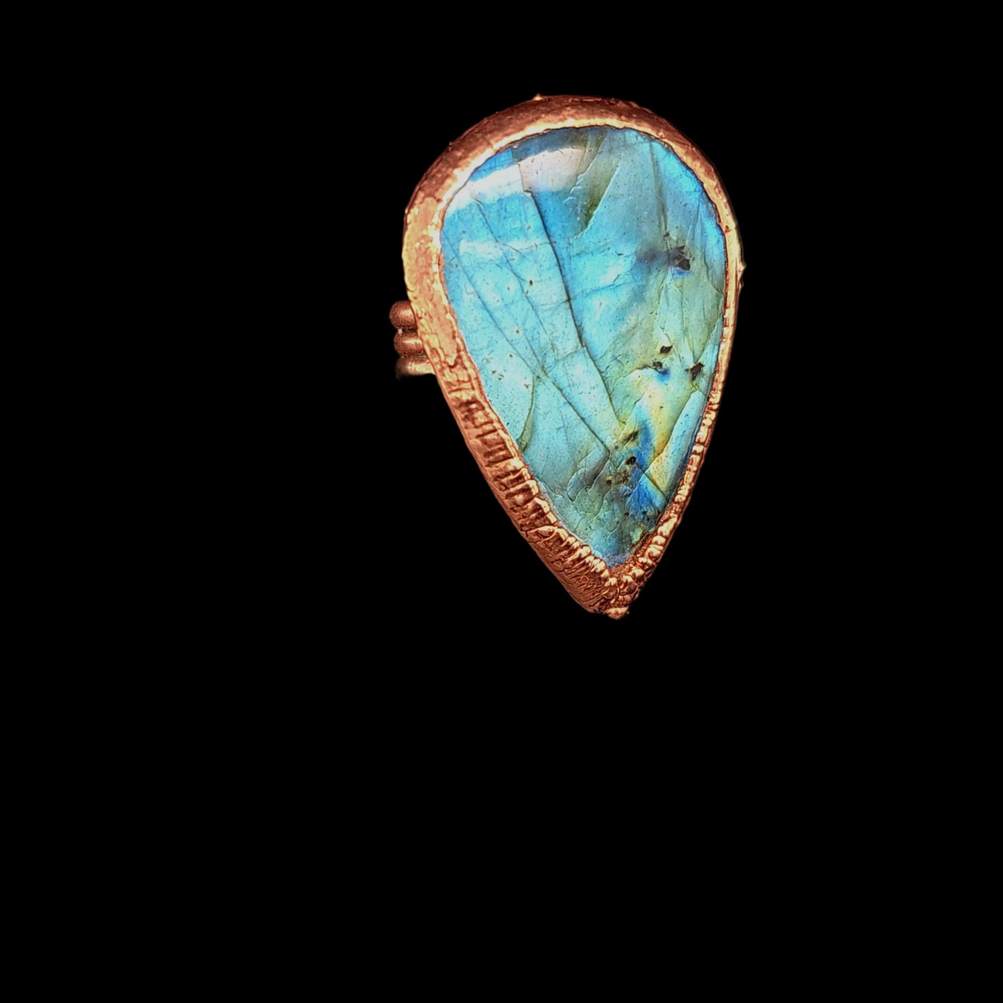 A teardrop-shaped labradorite ring with a textured copper-toned bezel, displayed on a black reflective surface. The labradorite stone exhibits vibrant blue and green iridescence.