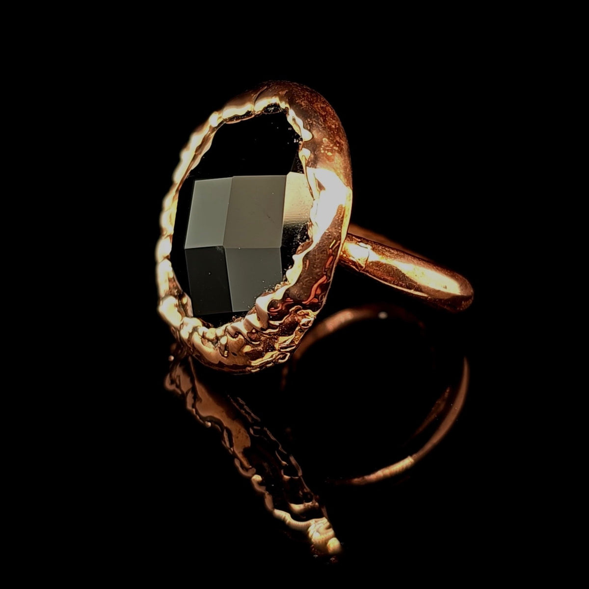 A copper ring with a large, irregularly shaped faceted black gemstone, photographed on a solid black reflective surface.