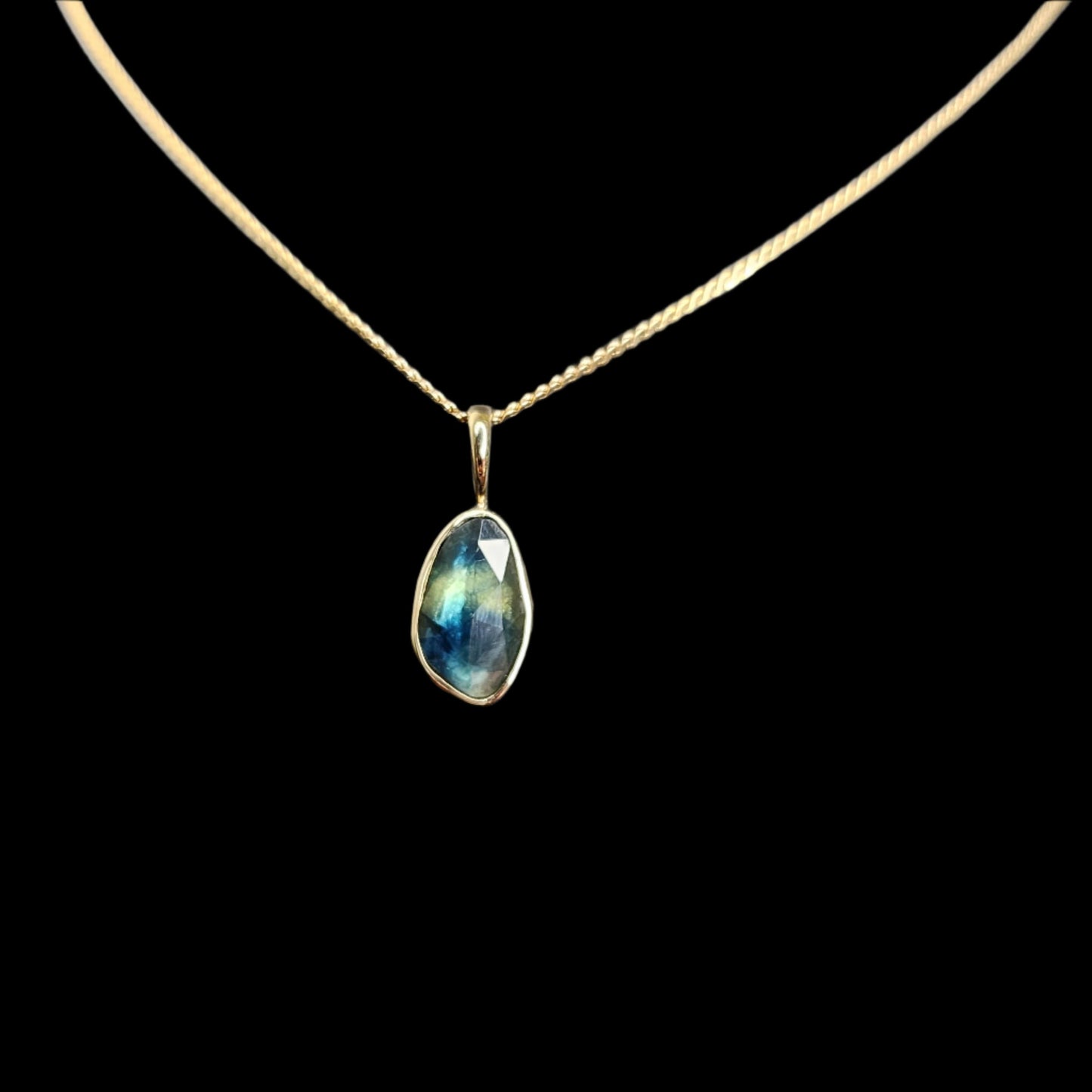 A delicate pendant featuring a rose-cut bi-color sapphire in shades of blue and green. The gemstone is set in a 14kt gold bezel with a matching gold bail, displayed on a simple chain against a black background.