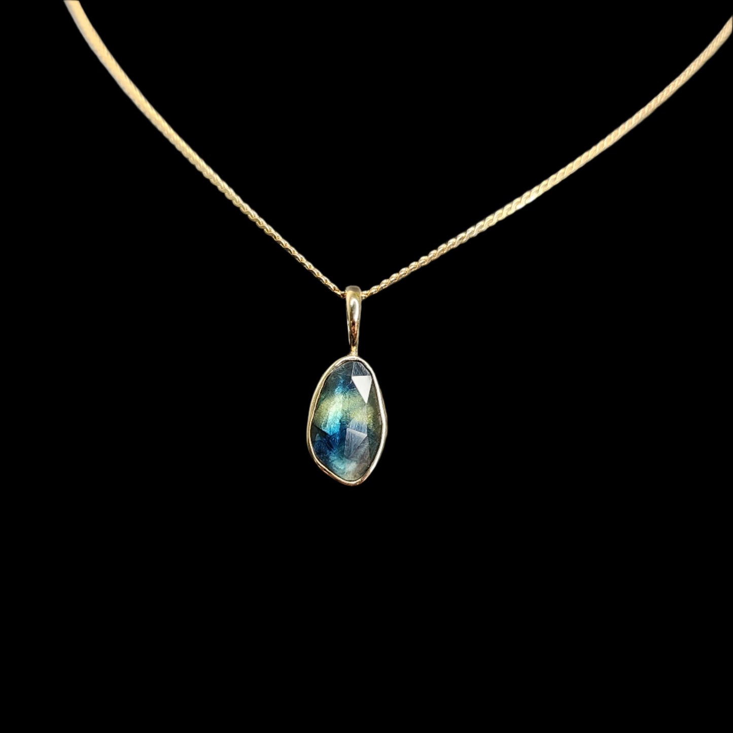 A delicate pendant featuring a rose-cut bi-color sapphire in shades of blue and green. The gemstone is set in a 14kt gold bezel with a matching gold bail, displayed on a simple chain against a black background.