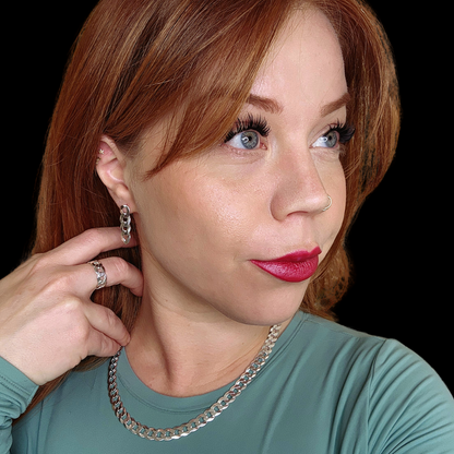 Portrait of a person with auburn hair wearing silver jewelry, including a curb link chain necklace, matching chain earrings, and a chain ring. They have red lipstick, bold eyelashes, and are dressed in a teal top with a black background