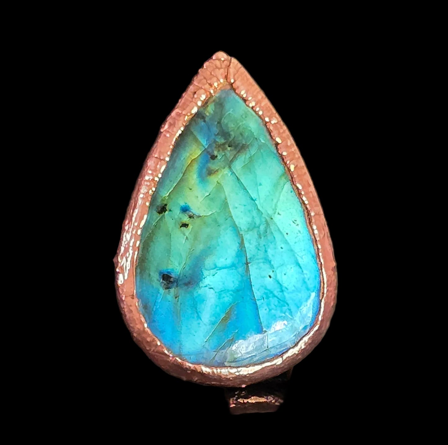 A teardrop-shaped labradorite ring with a vibrant blue and green iridescent stone set in a textured copper bezel, shown on a dark reflective surface.