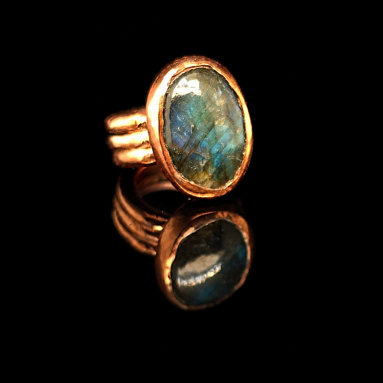 A copper ring with a large oval labradorite stone showing blue and green flashes, displayed on a reflective black surface.