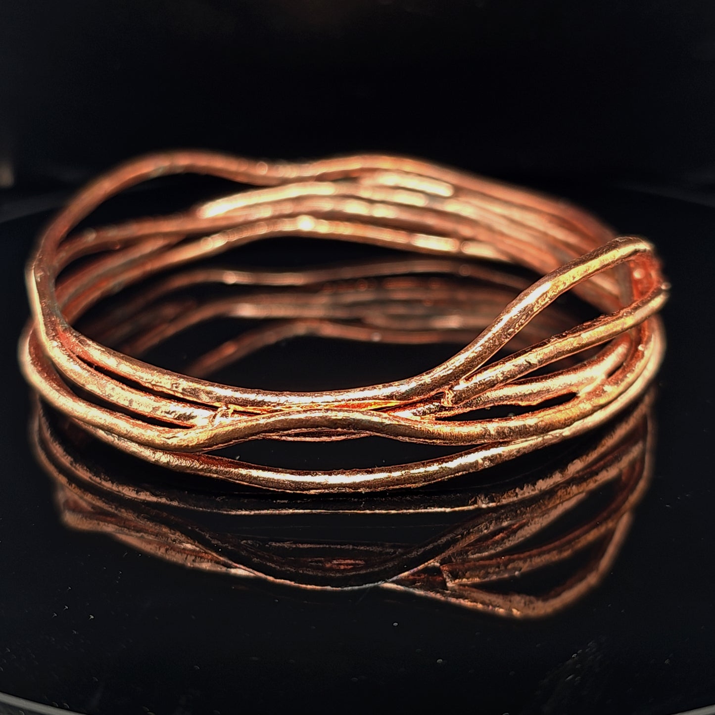 A copper-colored wavy wire bangle bracelet with a textured, organic finish, displayed on a glossy black reflective surface.