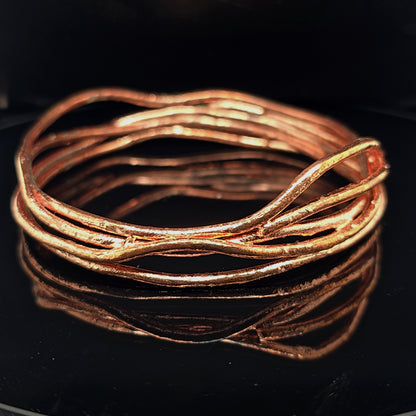 A copper-colored wavy wire bangle bracelet with a textured, organic finish, displayed on a glossy black reflective surface.