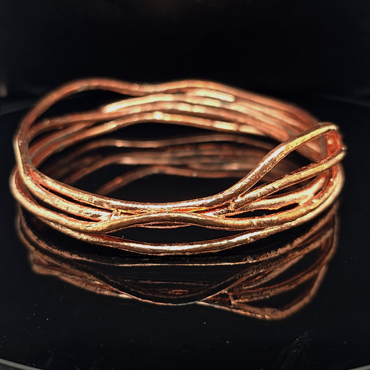 A copper-colored wavy wire bangle bracelet with a textured, organic finish, displayed on a glossy black reflective surface.