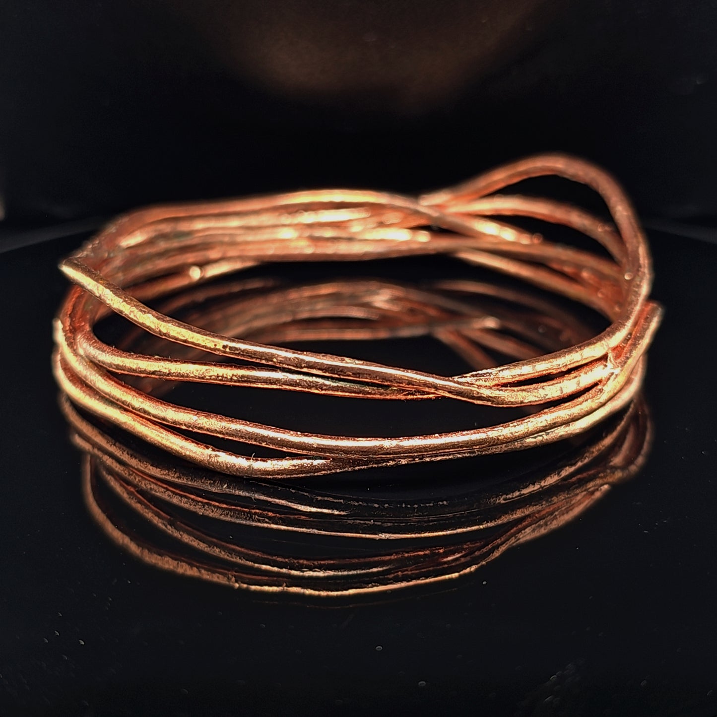 A multi-strand copper bangle bracelet with a textured, organic wavy design, displayed on a reflective black surface.