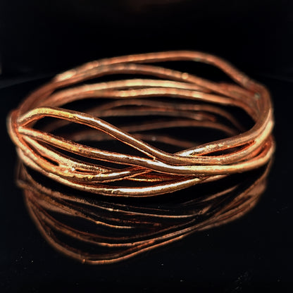 A hammered copper-toned metal bangle bracelet with a twisted, layered design, displayed on a reflective black surface.