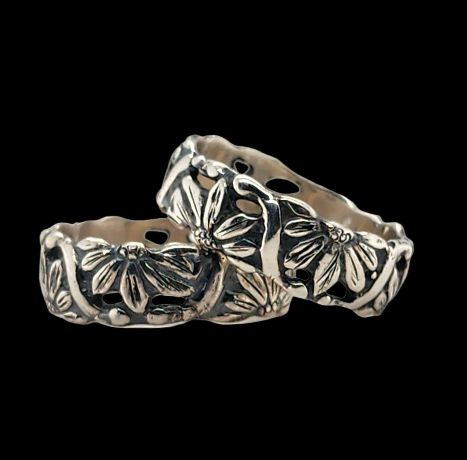 pair of silver floral-patterned ring, pattern goes all the way around the band, with half of flowers alternating directions, representing the duality of life, the natural world & partnership on a black background