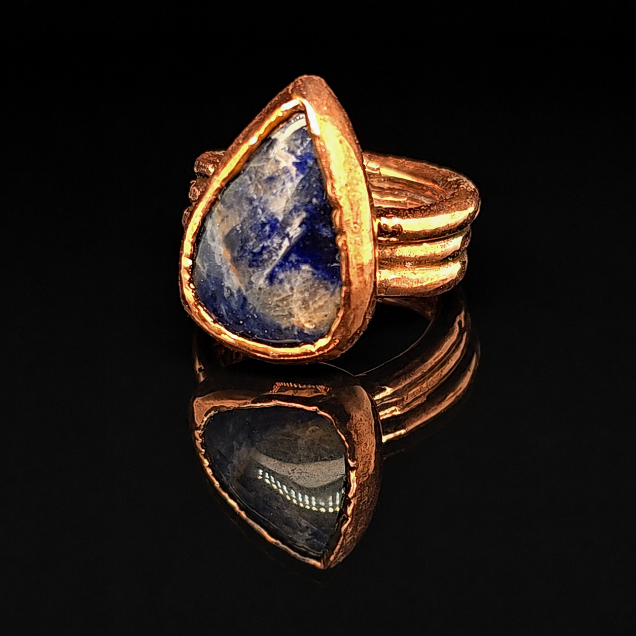 A teardrop-shaped ring featuring a natural blue and white stone set in a textured copper metal, displayed on a reflective black surface.
