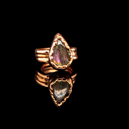 A teardrop-shaped ring featuring a natural iridescent labradorite gemstone in a textured rose gold setting, displayed on a reflective black surface.