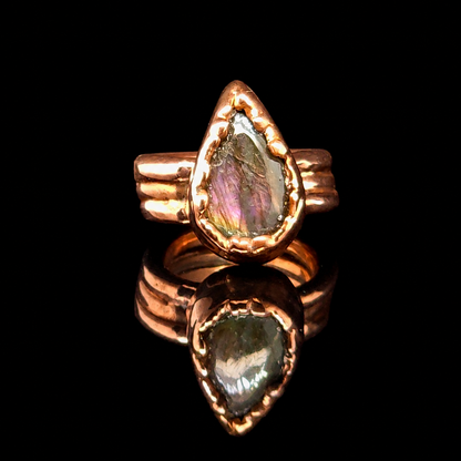 A teardrop-shaped iridescent gemstone ring set in a textured copper metal, displayed on a reflective black surface.