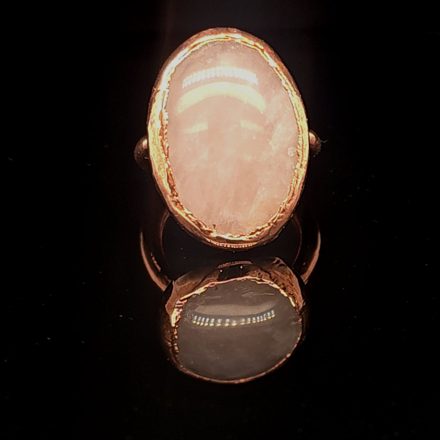 A copper ring featuring a large, oval cabochon gemstone with a pale pinkish-gold hue, displayed against a solid black background with a clear reflection below.