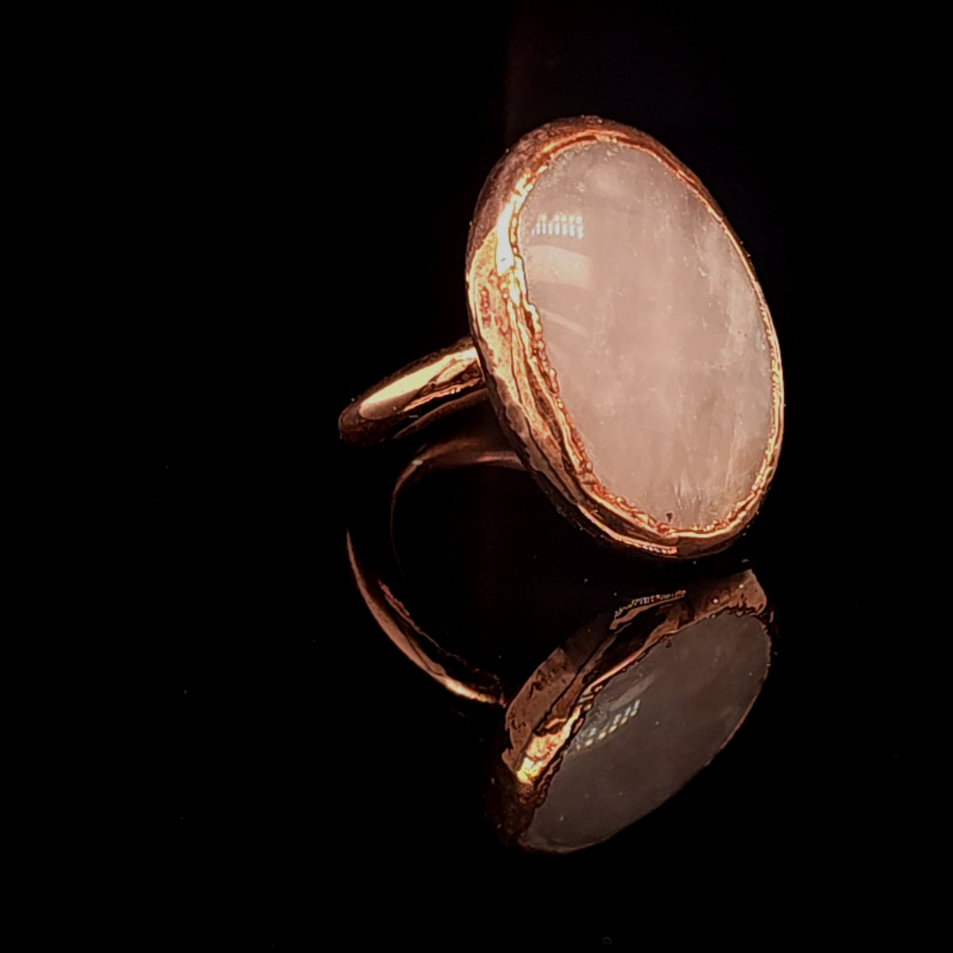 A handmade ring featuring a large, irregularly shaped pale pink stone set in a copper bezel, displayed on a reflective black surface.
