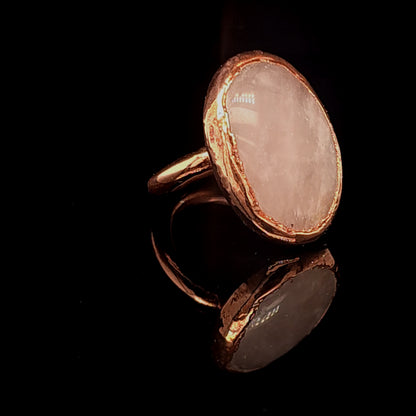 A handmade ring featuring a large, irregularly shaped pale pink stone set in a copper bezel, displayed on a reflective black surface.