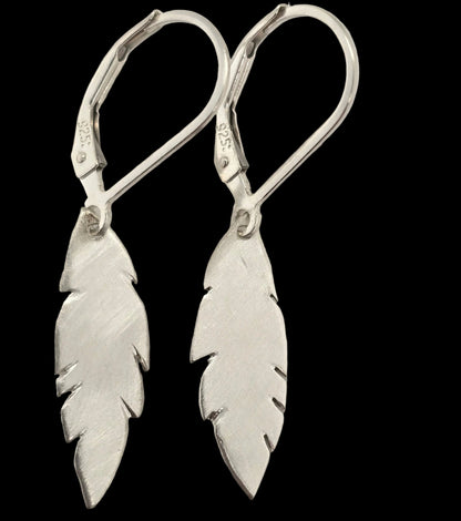 Pair of sterling silver feather drop earrings with a brushed finish, hanging from sterling silver leverback hooks on a black background.
