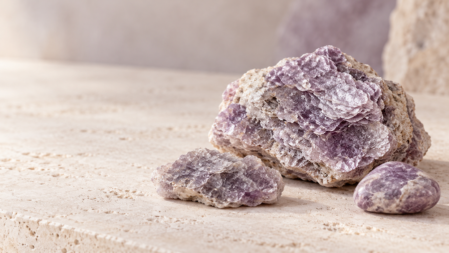 Raw lavender-purple lepidolite specimens with layered mica texture and a polished stone, arranged on a neutral travertine surface with soft lighting.
