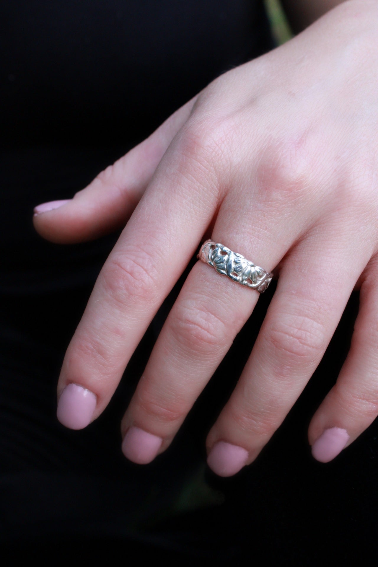 Close-up of a hand wearing a sterling silver floral ring with intricate flower and leaf engravings, displayed against a dark background.