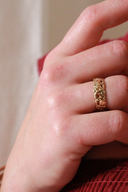 A close-up image of a hand wearing a gold ring with a detailed floral pattern. The hand rests naturally, and the background is softly blurred, featuring warm tones and subtle hints of fabric texture.