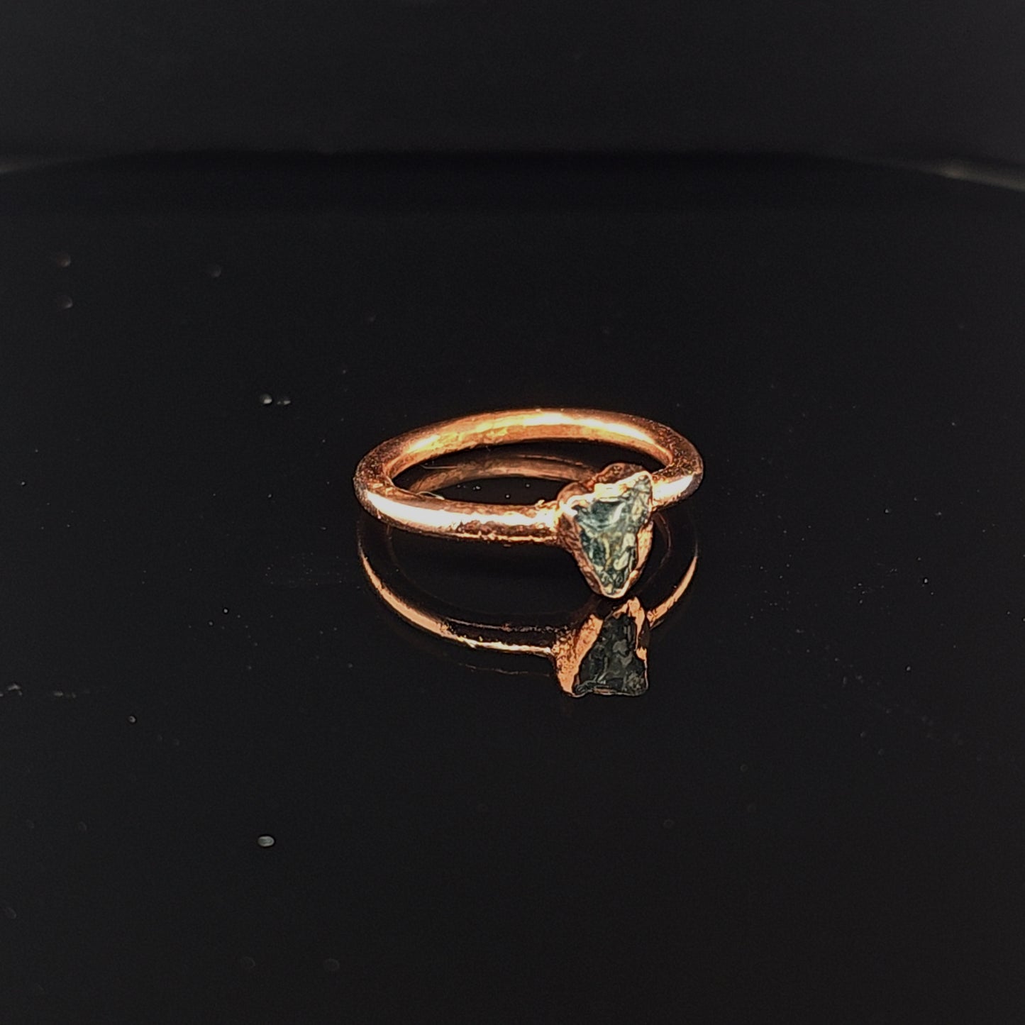 A rustic copper ring featuring a triangular teal apatite stone, displayed on a reflective black surface.