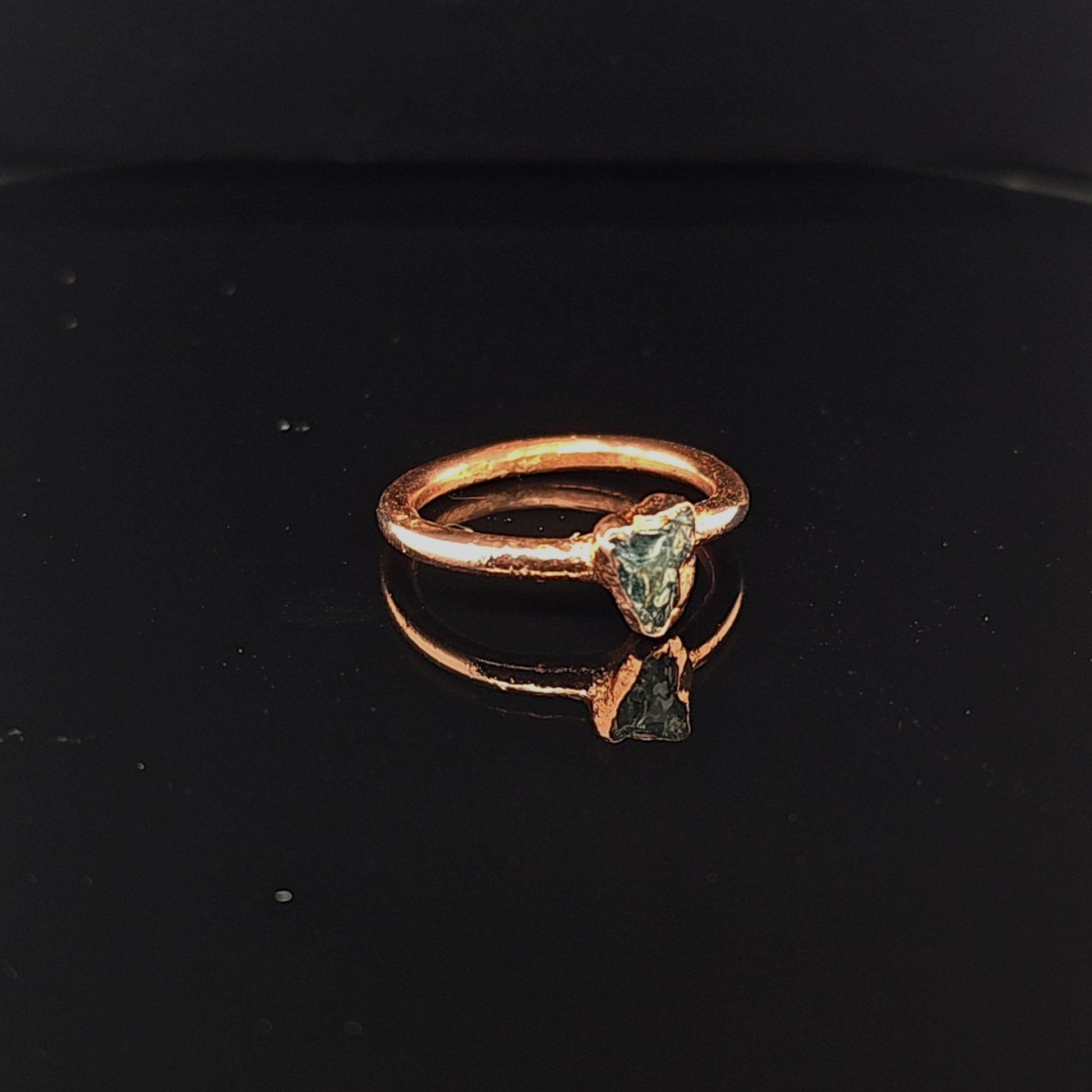 A rustic copper ring featuring a triangular teal apatite stone, displayed on a reflective black surface.