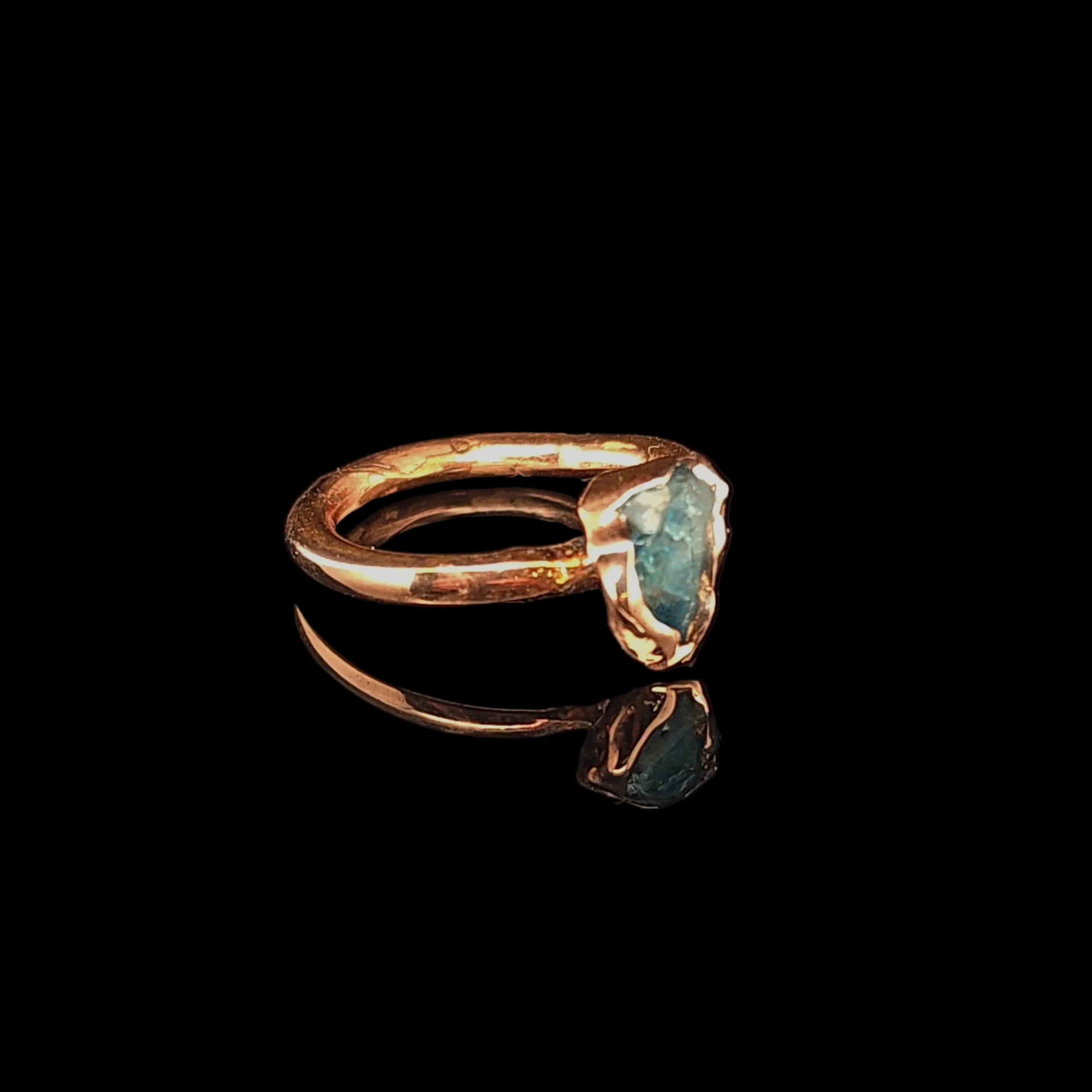 A handmade ring with a textured copper band and a raw, irregularly shaped blue-green gemstone, displayed on a reflective black surface.