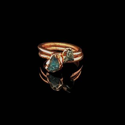 Two stacking rings featuring polished copper bands with two rough, triangular blue-green gemstones, displayed on a reflective black surface.