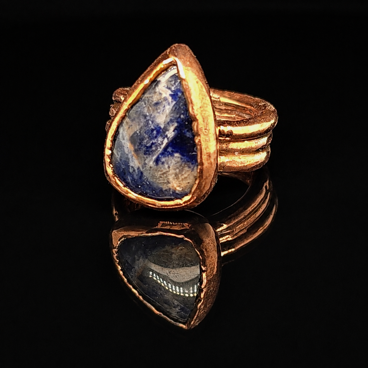 A copper ring featuring a large, teardrop-shaped blue peach sodalite stone, displayed on a reflective black surface.