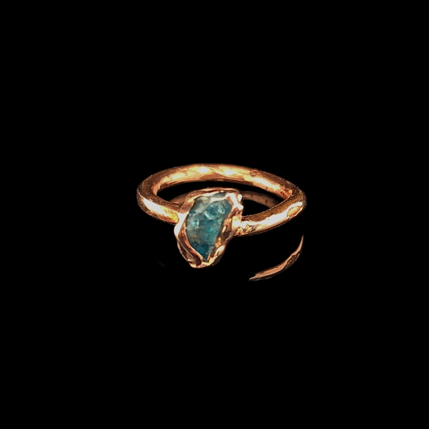 A handmade ring with a textured copper band and a raw, irregularly shaped blue-green gemstone, displayed on a reflective black surface.