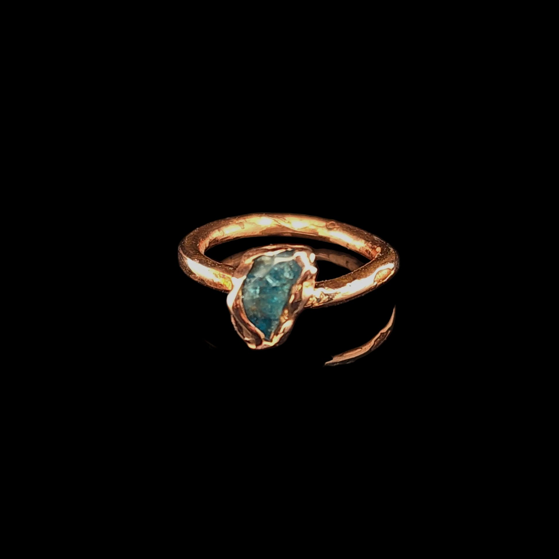 A handmade ring with a textured copper band and a raw, irregularly shaped blue-green gemstone, displayed on a reflective black surface.
