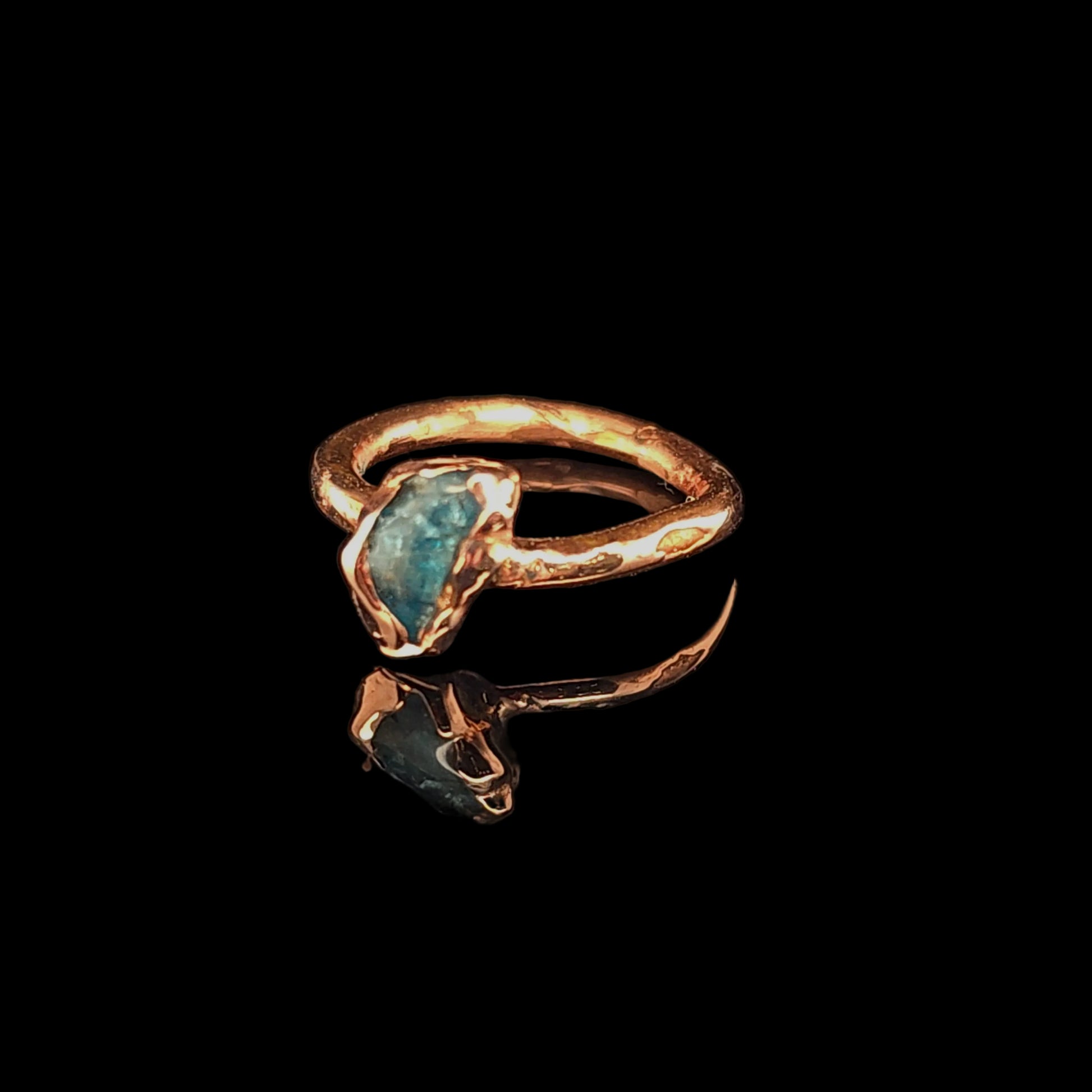 A handmade ring with a textured copper band and a raw, irregularly shaped blue-green gemstone, displayed on a reflective black surface.